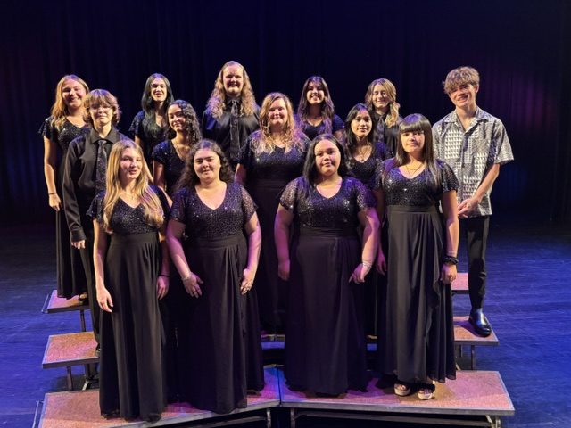 Members of the Kūlanihākoʻi Concert Choir posed in three rows on stage risers, wearing black formal attire with sparkled bodices.