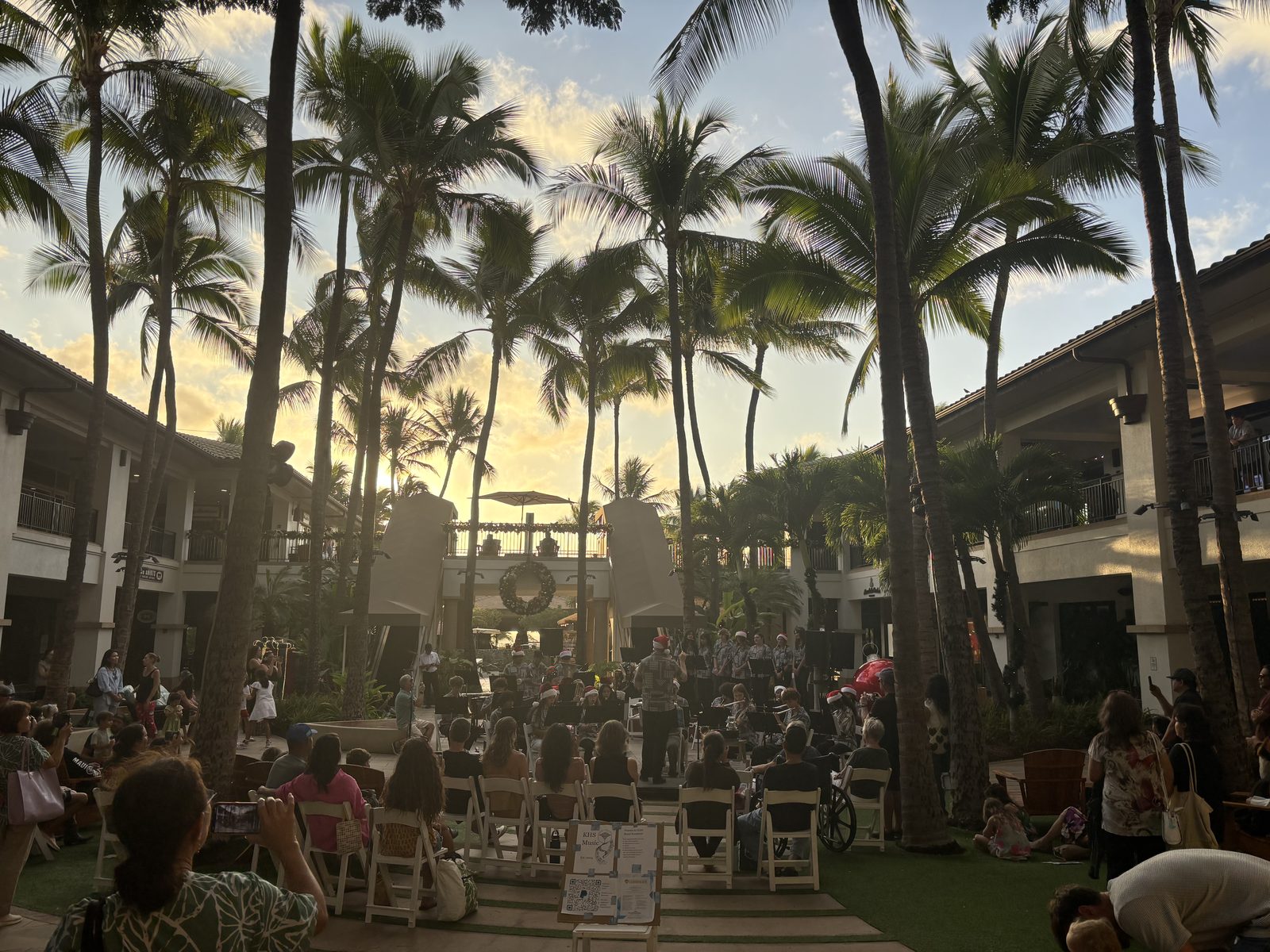 Students in Santa hats performing beneath palm trees as the sun sets over an open-air courtyard, with a seated audience watching.